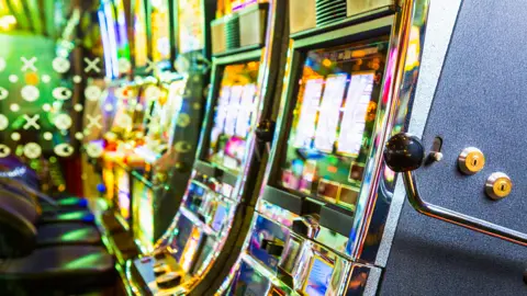 A close-up of a row of brightly lit slot machines. The machines are sitting side‑by‑side in a tight line, their reflective chrome edges catching the ambient neon.