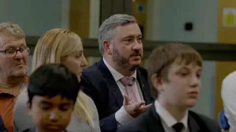 Channel 4 John Player is sat down speaking to a room as part of an audience. He is gesticulating with his hands and a number of people and students are around him. 