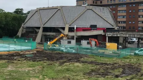 Reading Borough Council Hexagon theatre in Reading, with metal fencing around to secure the area for building. The image shows two diggers starting to dig up the earth around the theatre to install ground source heat pumps.