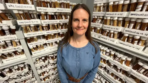 A woman with long dark hair and wearing a blue shirt standing in front of an L-shaped set of shelves piled high with Falcon tubes, containing substances that are varying shades of browns and beiges.