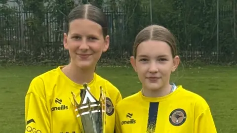 Nicole and Lexi stand side-by-side on the pitch in their yellow football kit holding a large silver trophy between them