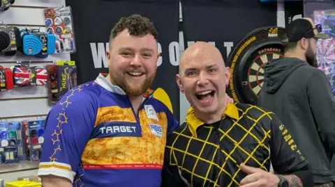 Brian Doran Two men standing side-by-side. One has Short dark curly hair and ginger facial hair. He is wearing a blue sports top with patterns. Next to him is a man with a bald head and a black top with a yellow pattern. Behind them are darts accessories.