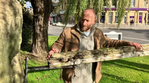 LDRS A man tends to a large wooden gate which appears rotten. The wood is attached to a metal black post