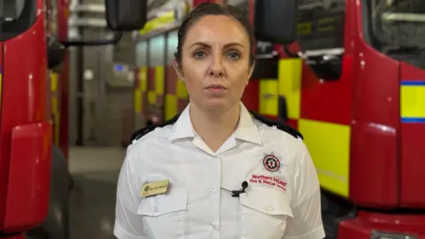 Mary-Kate McKevitt is standing between two fire engines, dressed in her uniform - white shirt, name badge, black epaulettes and the NIFRS logo. She has her dark hair tied back. She has blue eyes and she has her lips pursed.
