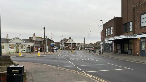 George Carden/BBC South Street looking towards Lancing railway station, there's a junction veering to the left. A tall brown building on the right, and the crossing can be seen in the distance. The road and pavements are almost completely empty