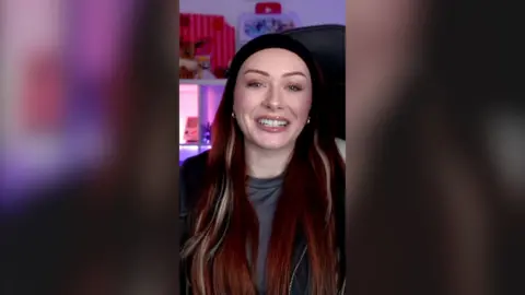 Elz The Witch has long red hair and is smiling at the camera