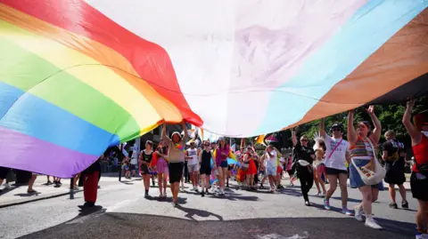 PA Media A shot taken looking underneath an enormous, parachute-sized Pride flag, looking down the parade at lots of people walking with smaller flags