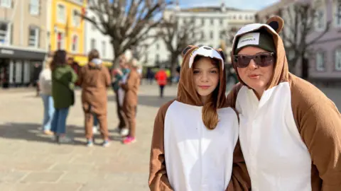 Sophie and Alexa look at the camera in their squirrel costumes. Sophie has a green beanie hat on and sunglasses. Alexa has long light brown hair which comes out of her costume. There's a group of people behind them some are dressed as squirrels. 