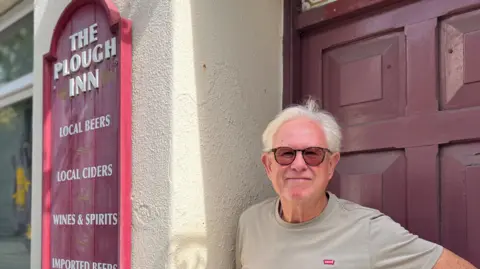 An man with grey hair, wearing a beige levi's t shirt and sunglasses. He stands in the doorway of a pub, next to a red sign with the "plough inn" and types of alcohol on it.