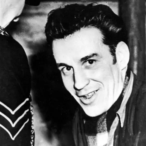 Getty Images Manuel, with thick dark hair and long sideburns, smiles at the camera. He is dressed in a dark jacket, open necked casual shirt and tartan scarf. A police sergeant is facing him, away from the camera.