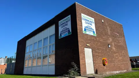 The brick building of Southwick Community Centre stands near the village green. The doors have been boarded up and are covered with metal sheets. 