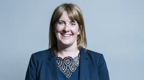 UK Parliament Sarah Jones pictured smiling against a white backdrop. She has short brown-blonde hair and wears a blue top underneath a black blazer.