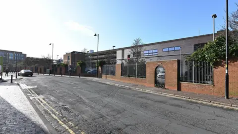 Hull City Council A road with an uneven-looking surface and faded double-yellow lines runs across the foreground, with a red-brick wall behind it. Large buildings stand in the background.