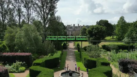 BBC Stately home gardens with a fountain and well pruned bushes. A monorail line is above with a green train travelling along it. 