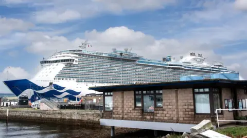 A large cruise ship is berthed at Invergordon. The ship has several floors and is blue and white in colour. There is an office in the foreground.