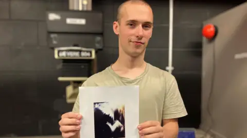 White male in his 20s with shaved head, wearing a t-shirt, standing in front of a laptop computer in a university dark room, holding a piece of paper in front of him
