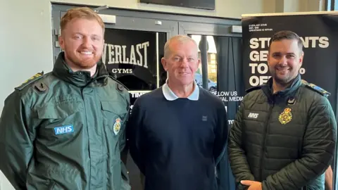 Richard Iles, in a blue jumper, stood between Jordan Griffiths and Lee McLaren, who are wearing their paramedic uniforms, looking at the camera while stood at the doors of the gym