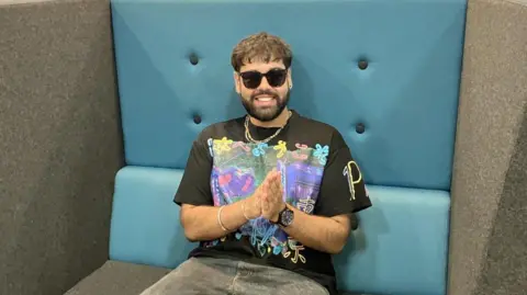 A mean wearing dark sunglasses inside, a colouful t shirt and chains around his neck sits in a huge blue and grey chair
