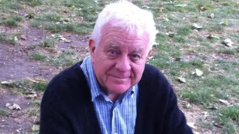 Geoff, de la Policía de Cambridgeshire, es un hombre de pelo blanco. Lleva una camisa azul a cuadros y un cárdigan azul marino. Está sentado en el césped, sonriendo a la cámara.