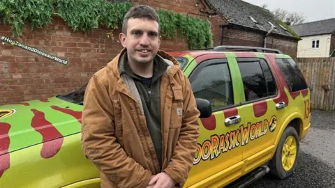 A man wearing a brown jacket stands in front of a yellow, green and red car with a brick wall and buildings behind him