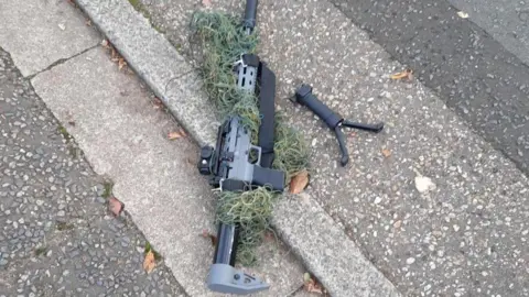 A grey and black air rifle placed on a concrete road. The rifle handle is detached from the gun, loose on the ground.