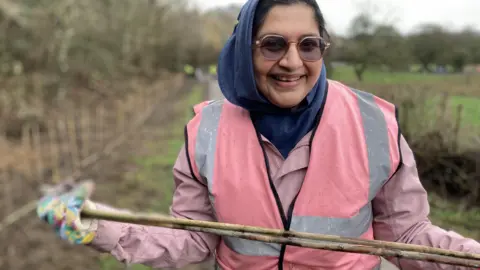 A woman in a pink high vis jacket and rain coat wearing a blue hijab smiles at the camera