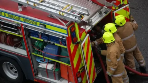 BBC Three firefighters in full safety clothing and helmets link a water hose to the back of a fire engine