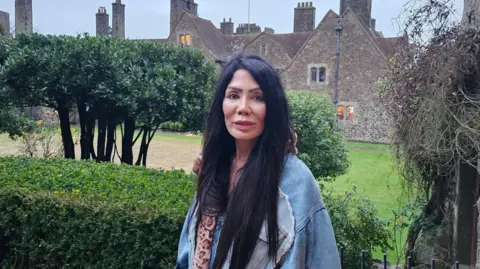 Ann Kaplan Mulholland standing in front of the west wing of Lympne Castle. She has long black hair and a blue jacket on. She is looking directly at the camera.