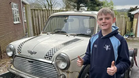 @Miniboysfirstcar A 10 year old boy standing in front of an old cream-coloured mini with rust visable.