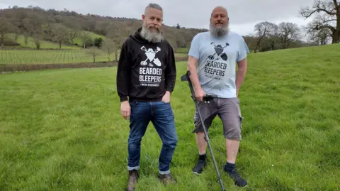 Two men with a metal detector in a field of green grass. 
