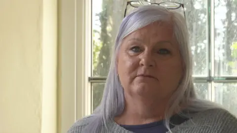 Tyler's key worker, Alex, has white hair with a purple streak in it. She is wearing a grey jumper and sits in front of a window in a yellow-painted room. 