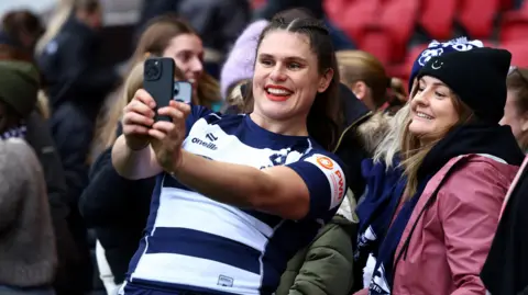 Bristol Bears' Ilona Maher takes a selfie with fans after the match with Gloucester/Hartbury at Ashton Gate. She is smiling as she holds the phone out to take the picture, as are the female fans in the background