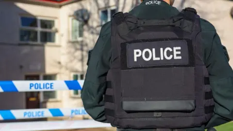 The torso of a police officer seen from behind hearing a bullet-proof vest with the word "Police" on the back. In front of them an apartment block surrounded by blue and white police tape.