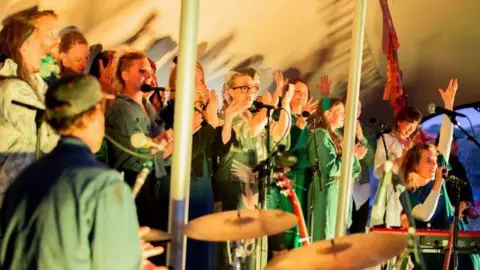 Oak Choir A group of people singing, some with their hands in the air and a person on a drum kit near the camera.