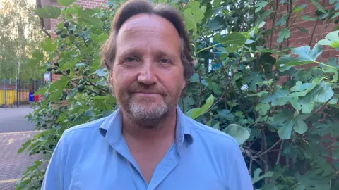 A man is standing outside a council building, wearing a blue shirt with a collar - behind him is a large green bush. 