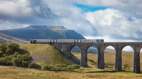 A Northern train crossing the Ribblehead Viaduct. The stone structure has several arches and is surrounded by open green space, with a fell behind it.