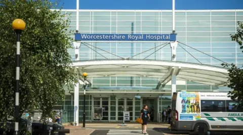 Worcestershire Royal Hospital A generic image outside Worcestershire Royal Hospital. There is a silver ambulance parked outside and a woman walking across the zebra crossing holding a leather bag and wearing a black dress with a lanyard on top.

