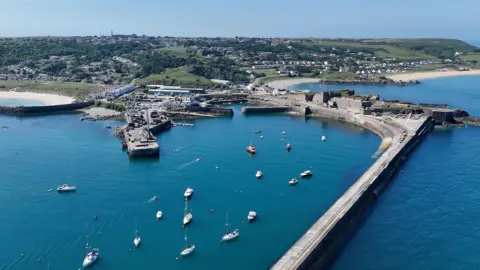 BBC Alderney's harbour on a clear day. There are boats moored behind a sea wall. Behind the harbour are green fields and housing.