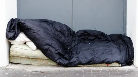 A dirty mattress on the floor outside double grey doors. There is a black sleeping bag laid on top of the mattress.