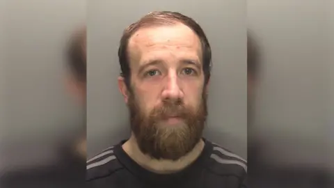 Police custody image of Lee Merrison he has short brown hair and a beard and is wearing a dark shirt with grey stripes on the shoulders