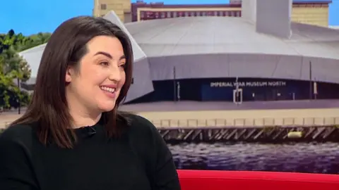 Bianca Perea, wearing a black jumper, has long dark hair. She smiles as she is being interviewed on BBC Breakfast's red sofa.