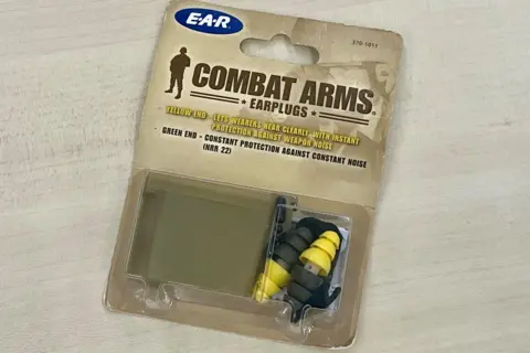 A packet of military earplugs in a box lies on a table. The box says Combat Arms Earplugs, with instructions on how to use them and what they do. A plastic box is in the packaging next to a set of the earplugs, which are yellow on one side and dark green on the other.