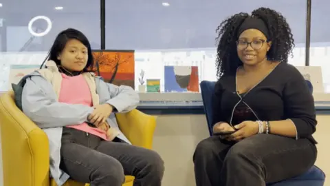  A woman in a pink shirt sits on a yellow chair and a woman in a black outfit sits on a navy blue chair