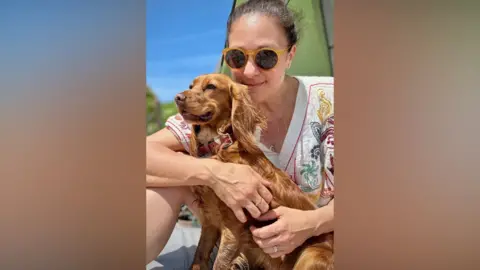 Shelley Perkins Shelley Perkins smiles at the camera as she holds her dog Roxy, a golden cocker spaniel. Shelley has dark brown hair and is wearing sunglasses and a floral top.