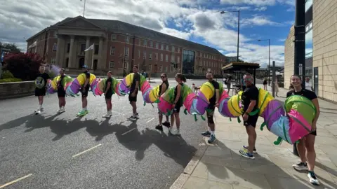 Amy Woolliscroft Picture of ten runners in a line ready to start the Ramathon, each joined by a multi-coloured tube creating a caterpillar costume in Derby city centre.