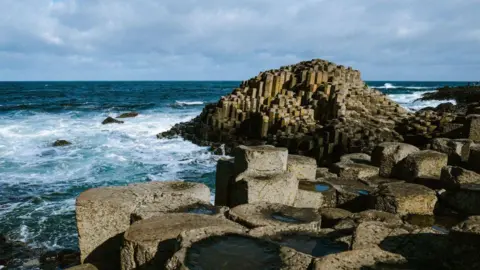 Hexagonal columns all fitted together at the edge of the coast. The sea is rough and has white foam at the rocks. 