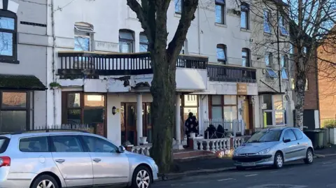 Laura Coffey/BBC A white building with a long brown balcony on its second floor with many glass windows. Two silver cars are parked outside the front of the building.