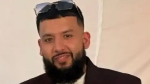 Police handout Syed is wearing sunglasses on top of his head and is smiling at the camera. He has a beard and is wearing a dark coat with a white top underneath.