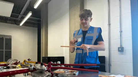 Emma Baugh/BBC Oliver is wearing a blue hi-vis vest with a name badge on. He is standing in a workshop and holding a metal pipe which he appears to be measuring the thickness of with a device.