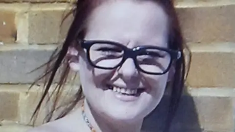 Family Handout A woman in dark glasses with dyed auburn hair smiling. She is standing in front of a brick wall.